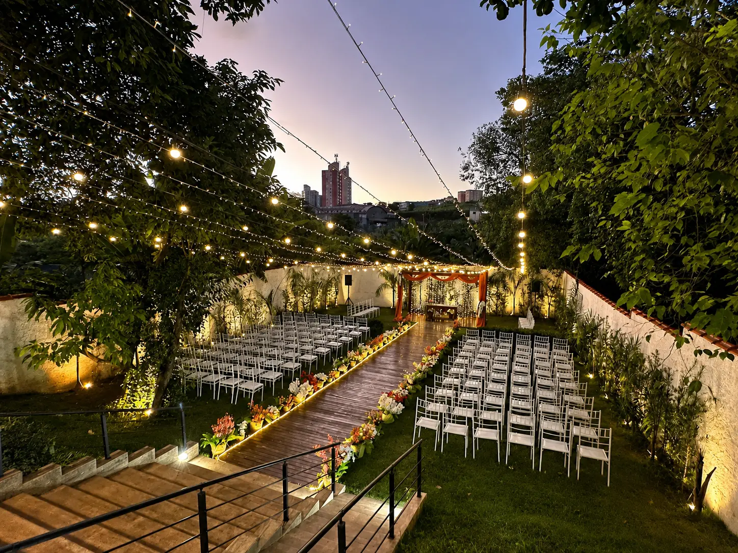 Espaço ao ar livre decorado para cerimônia de casamento ao entardecer, com cadeiras brancas, luzes suspensas, flores coloridas e um corredor central de madeira