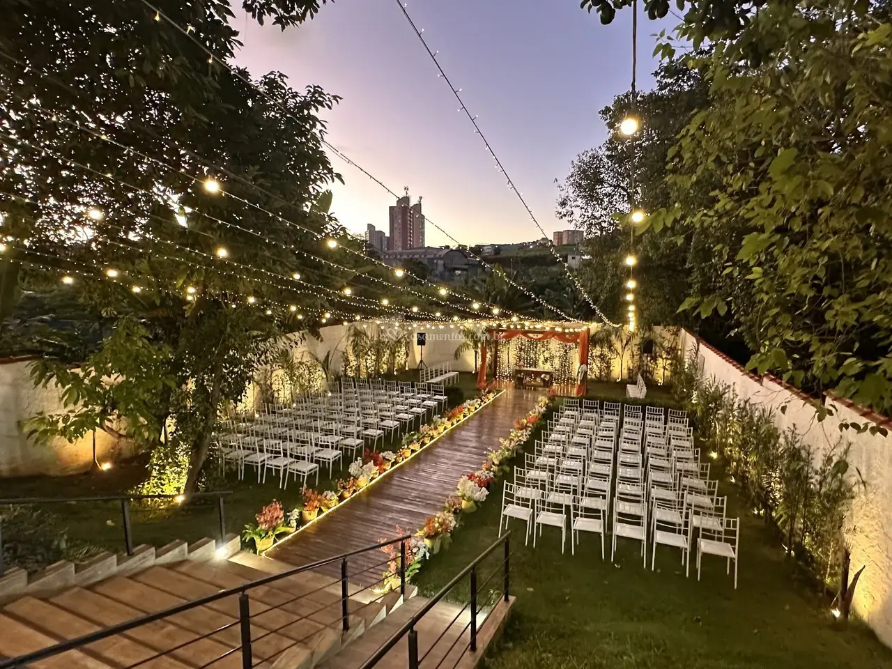 Espaço ao ar livre decorado para cerimônia de casamento, com cadeiras brancas, luzes suspensas e flores ao entardecer